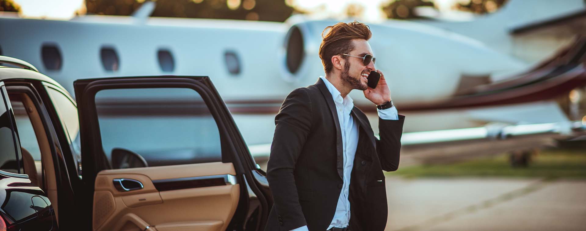 Smiling affluent man talking on the phone while getting out of a car with a private jet in the background