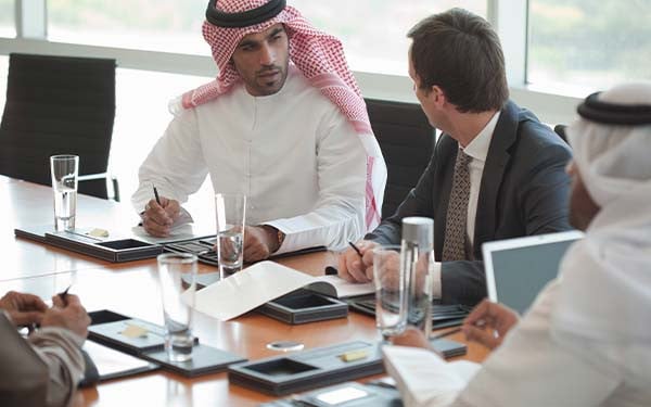 Three businessmen in discussion around a boardroom table, two in Arabic attire and one in a traditional suit