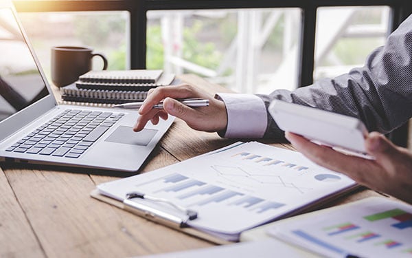 Closeup image of a person analyzing charts on a desk and scrolling on a laptop with a calculator in one hand